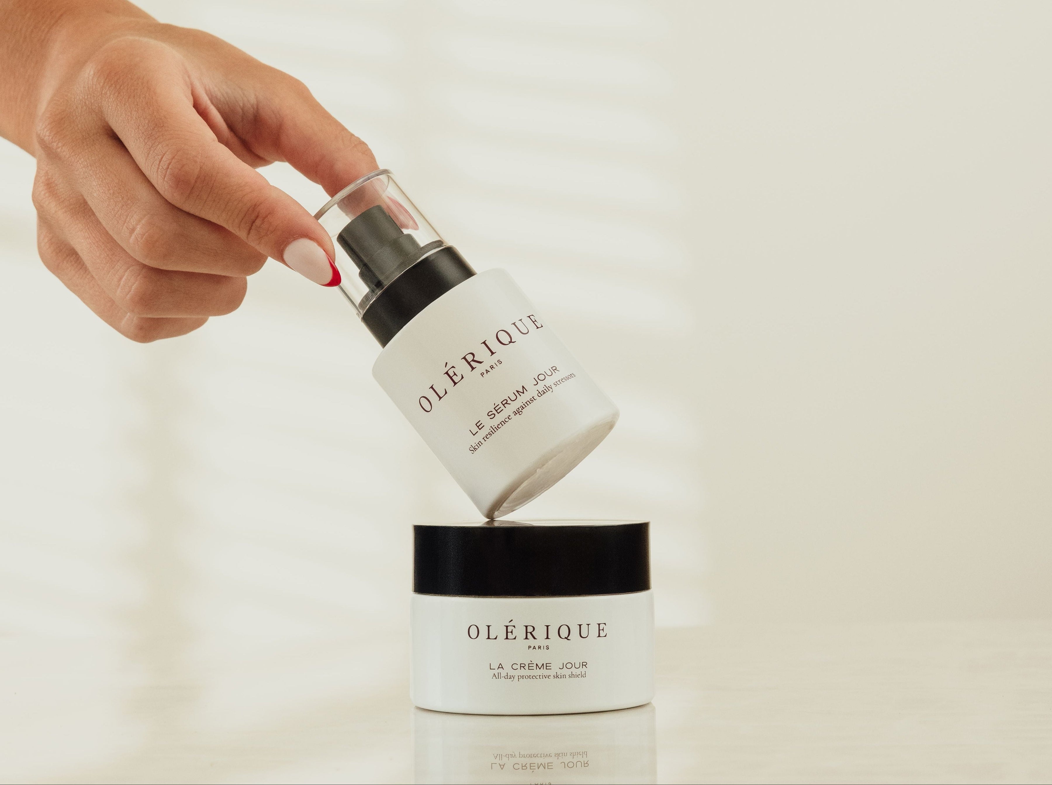 A hand holds a small white bottle above a matching jar labeled OLÉRIQUE, against a soft cream background with faint window-light shadows.